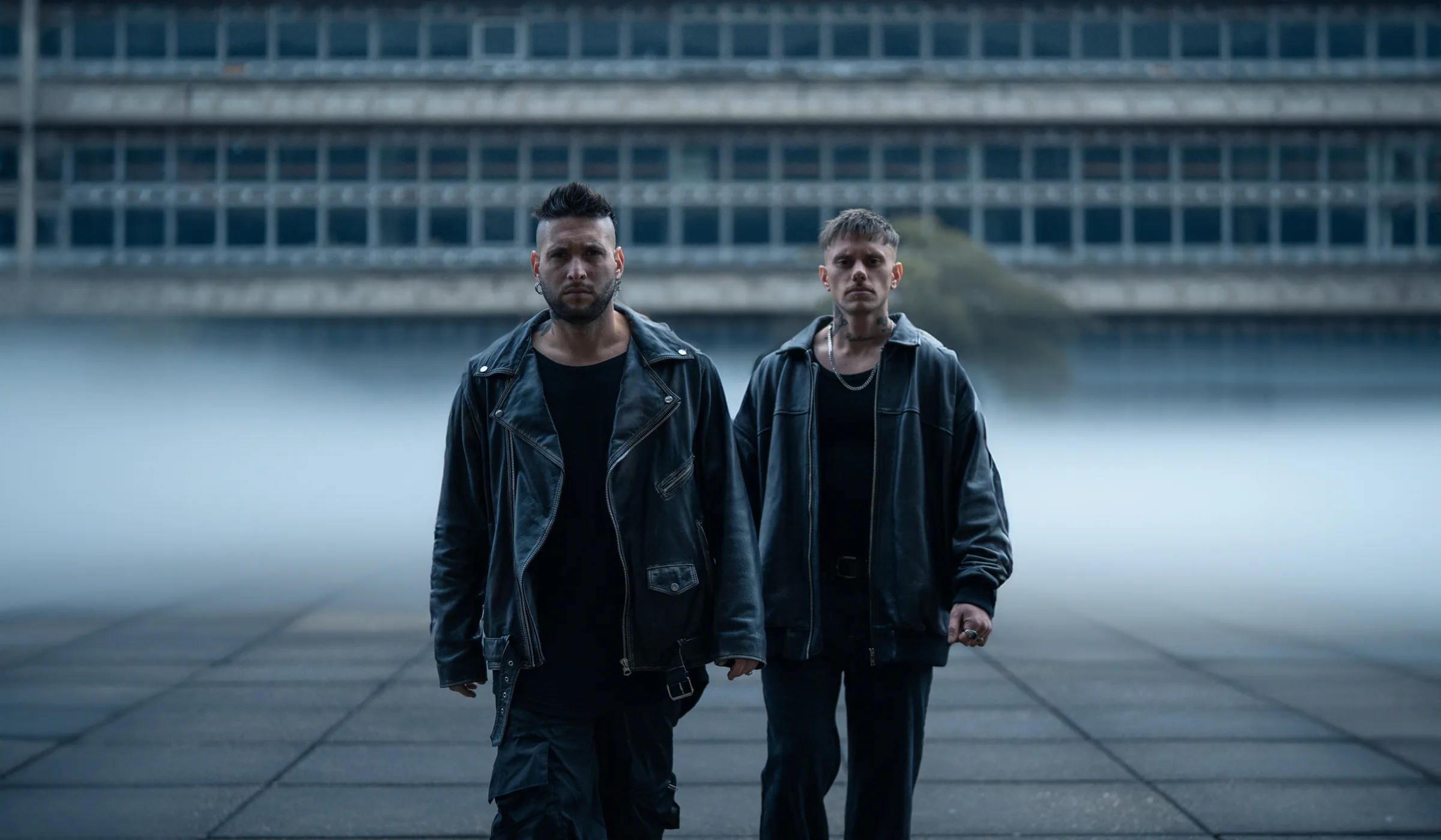 Mateo D'Angelo y Felipe Bonilla caminando frente a un edificio brutalista en la niebla